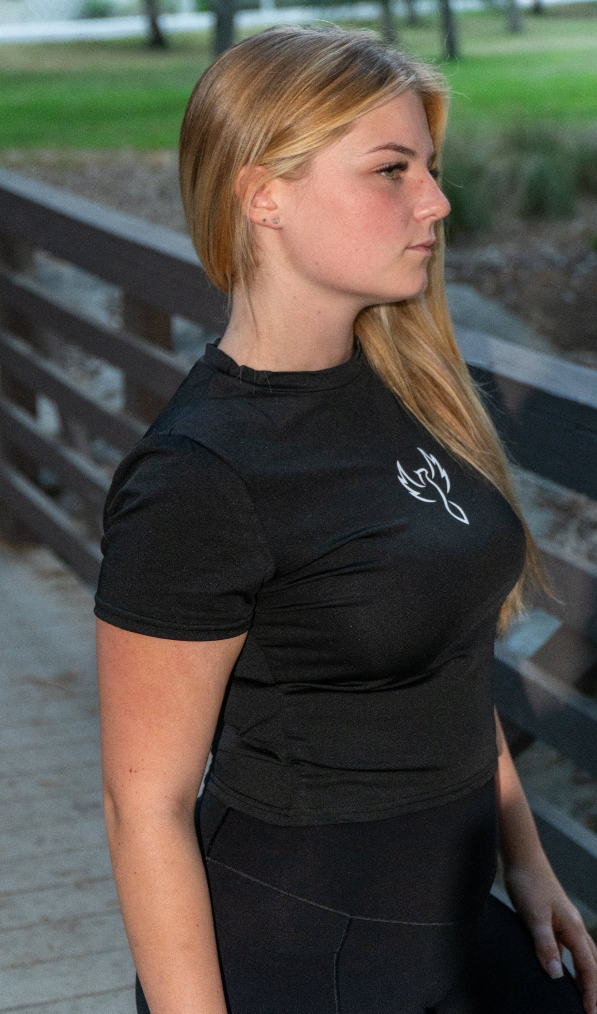 A woman with long blonde hair is shown in profile wearing a fitted black short-sleeve athletic top with a small white chest logo, standing outdoors on a wooden bridge.