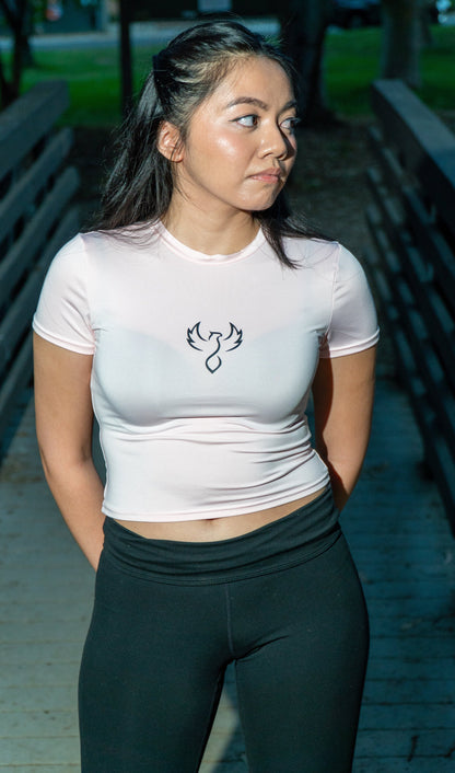 A woman wearing a fitted white short-sleeve athletic crop top with a black bird logo on the chest, paired with black leggings, standing outdoors on a wooden walkway.