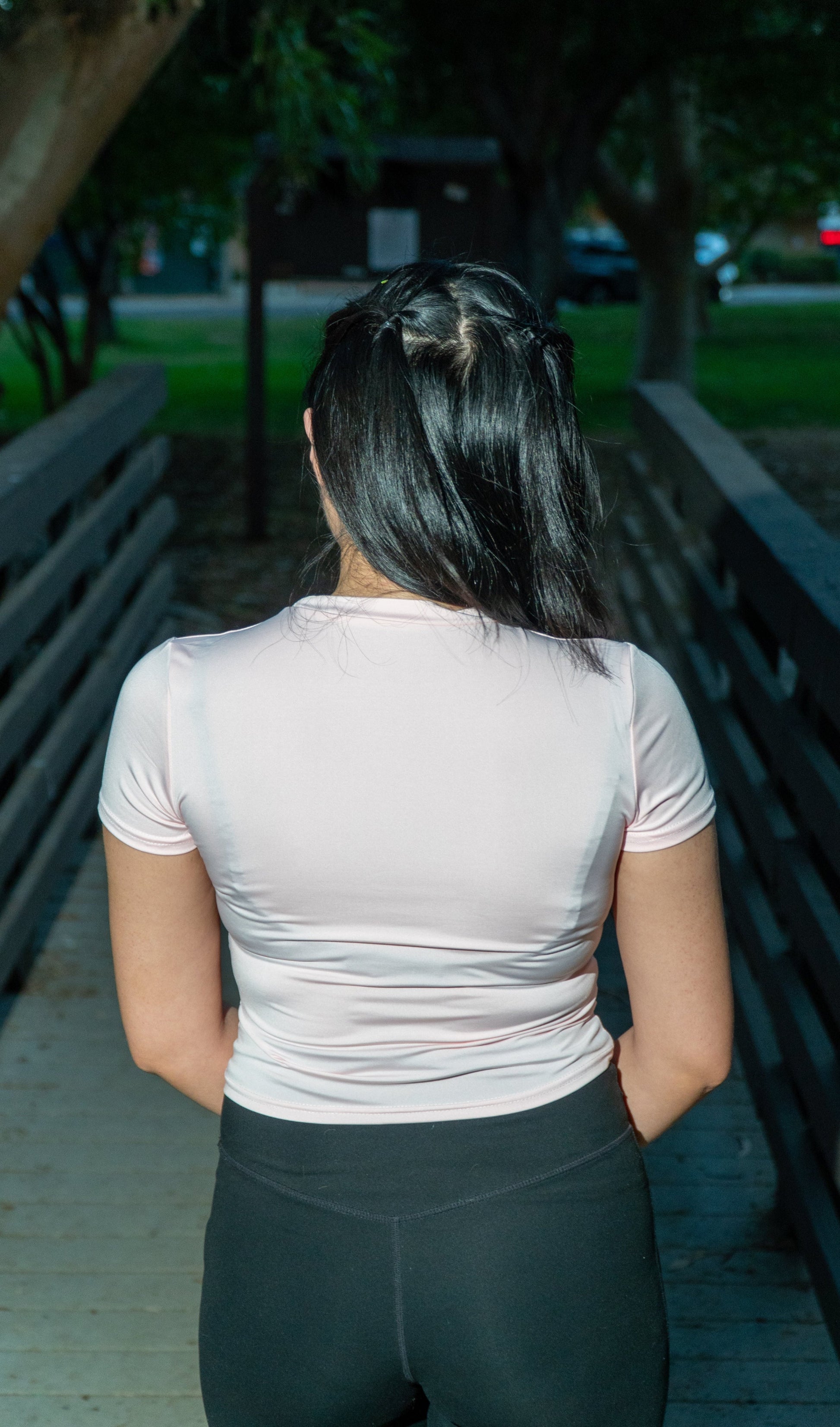 A woman viewed from behind wearing a light pink short-sleeve athletic top and black leggings, standing on a wooden boardwalk outdoors with trees and park seating in the background.